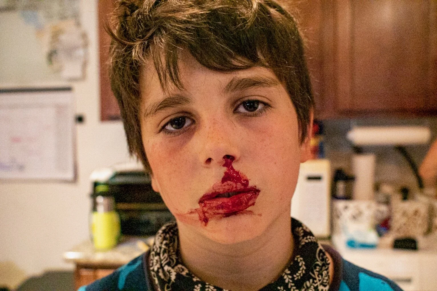 A boy with short brown hair and light skin, with makeup and fake blood on his face, looking directly at the camera in a kitchen setting.