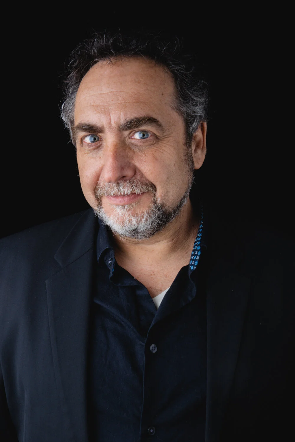 A middle-aged man with curly dark gray hair, a gray beard, and blue eyes. He is wearing a dark blazer over a dark shirt with a patterned inner collar, against a black background.