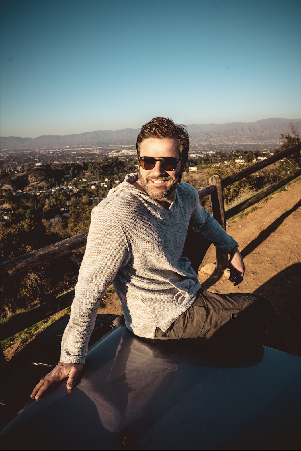 A man with sunglasses and a gray hoodie sitting on a car hood on a hill overlooking a city and mountains during sunset.