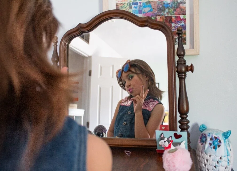 A young woman with brown hair and sunglasses on her head looking into a mirror, making a peace sign with her fingers and pouting her lips, in a bedroom with decorative items and colorful artwork.