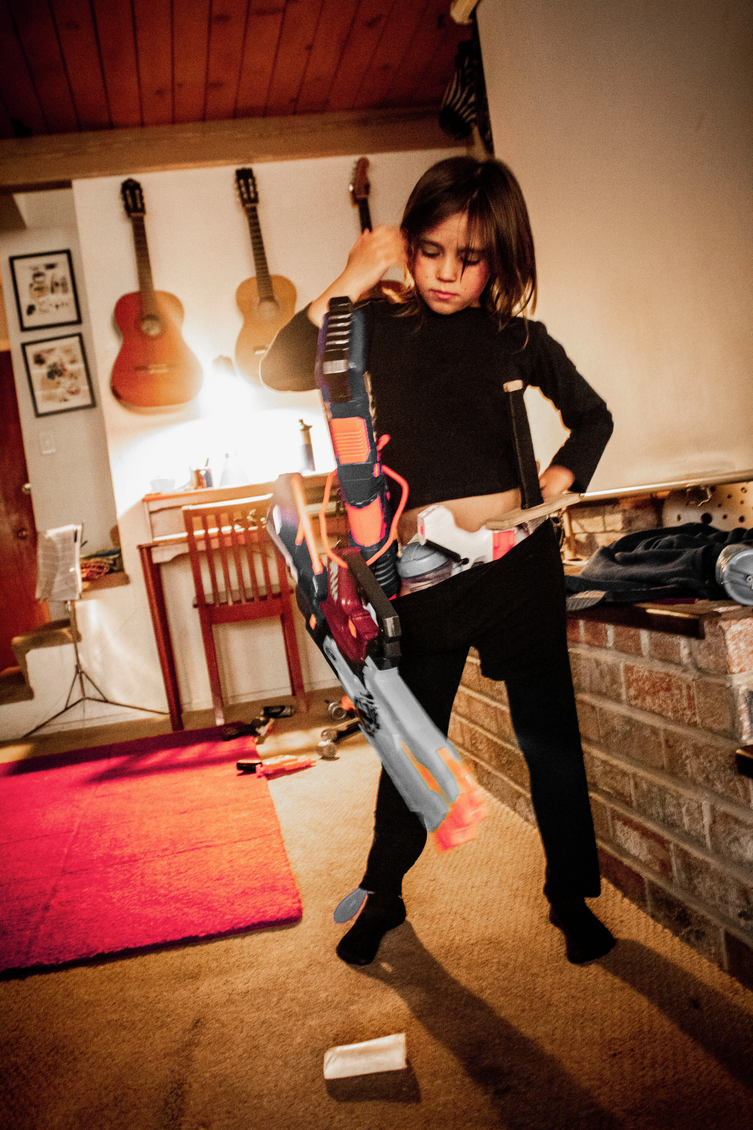 A young girl with dark hair and a black long-sleeve shirt holding a nerf gun inside a cozy room. The room has a brick fireplace on one side, decorated with hanging guitars on the wall, and a red rug on the carpeted floor.