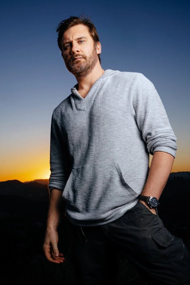 A man with brown hair and a beard wearing a gray hoodie and black pants poses outdoors at sunset, with a mountain landscape in the background.