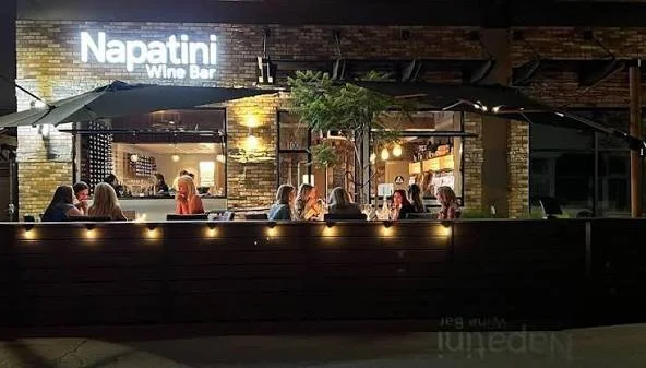 People dining outside at Napatini Wine Bar during nighttime, with outdoor seating, umbrellas, and warm lighting inside the venue.