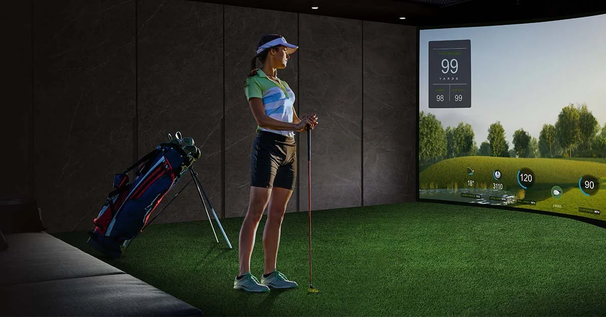 Woman at an indoor golf simulator, ready to hit a shot, with a golf bag on a stand beside her and a large screen displaying a golf course and statistical data about her shots.