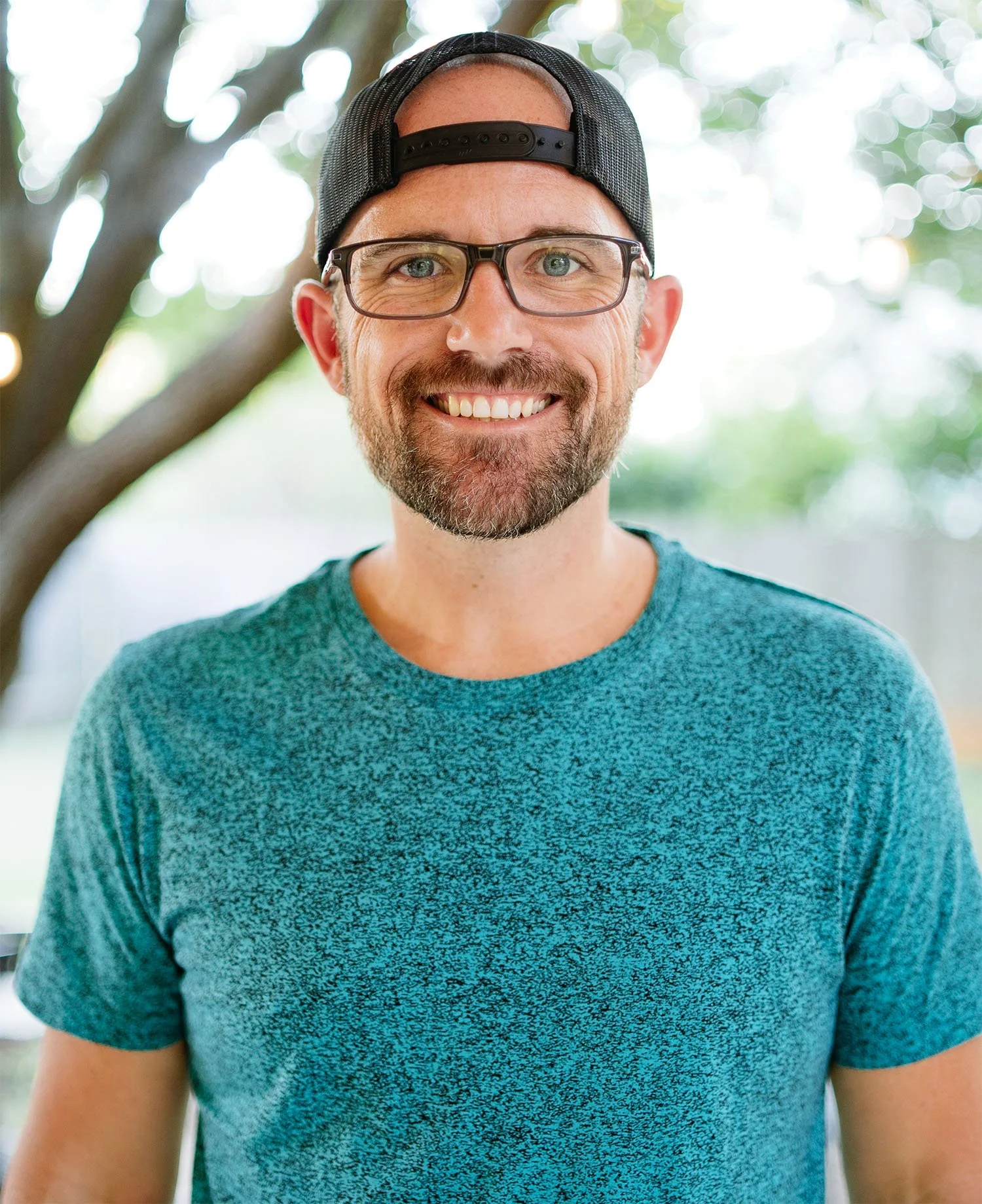A smiling man with glasses, a beard, and a mustache, wearing a black cap backward and a teal heathered T-shirt, standing outdoors with blurred trees and sunlight in the background.