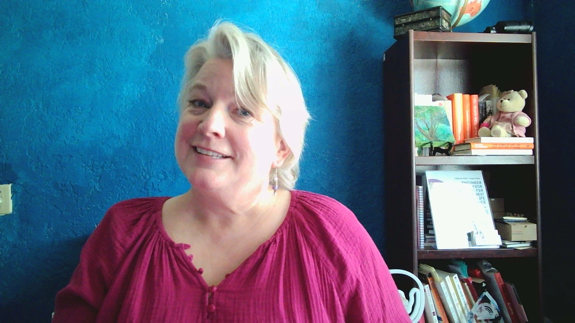 Jennifer Jank with short blonde hair wearing a pink blouse smiling in front of a blue textured wall with a bookshelf filled with books and decorative items behind her.