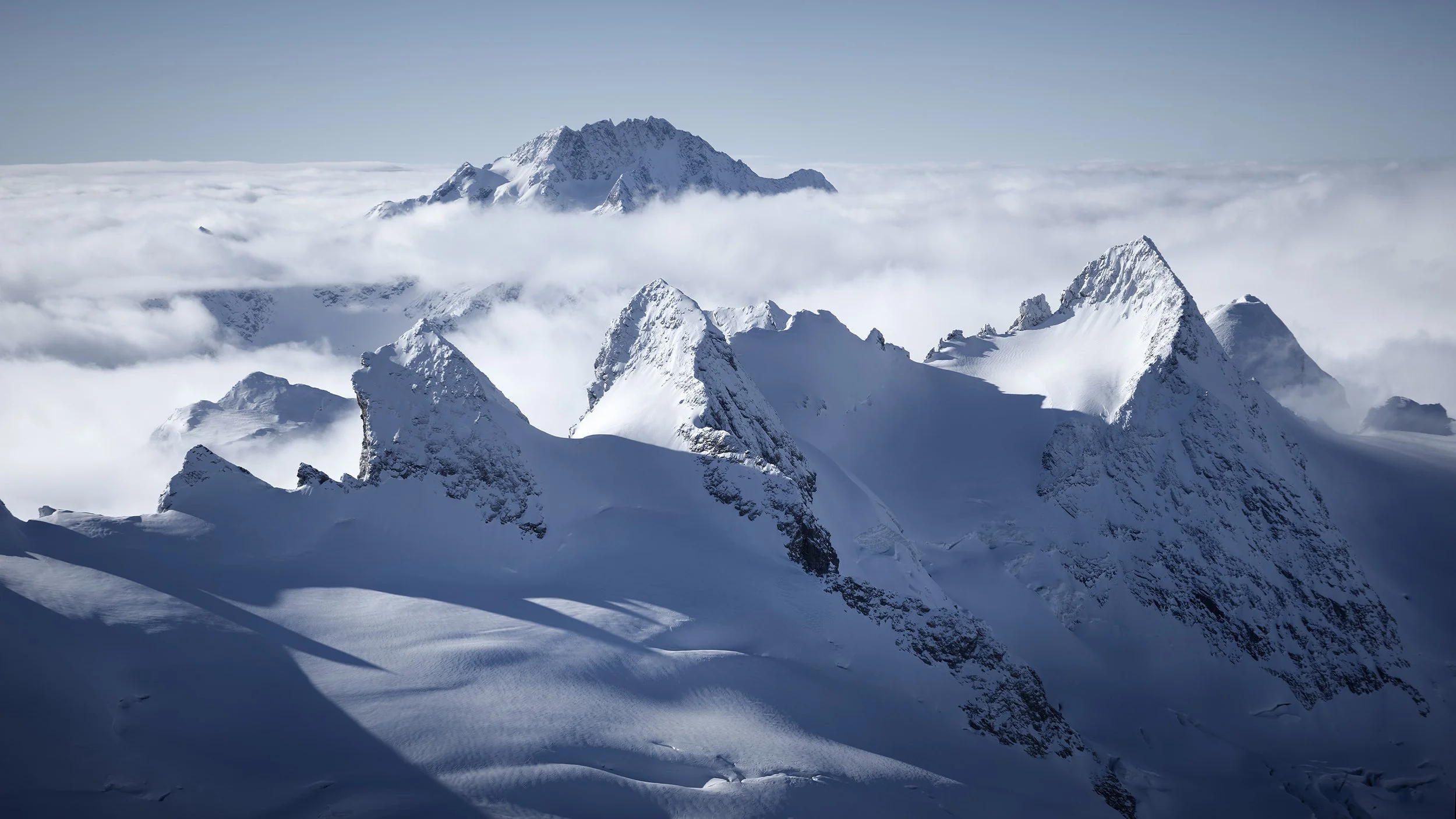 Aerial fine art photograph by Jürg Kaufmann showing La Sella and Piz Glüschaint rising above a sea of clouds in the Bernina Range, with Monte Disgrazia on the horizon, taken from a helicopter in December 2025 over the Engadin, Switzerland.