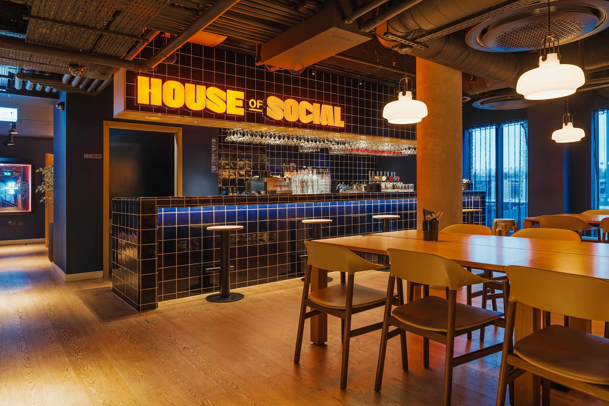 Interior of a bar and dining area with a sign saying "House of Social" behind the bar, which has dark blue tile, surrounded by wooden tables and chairs, and dim lighting.