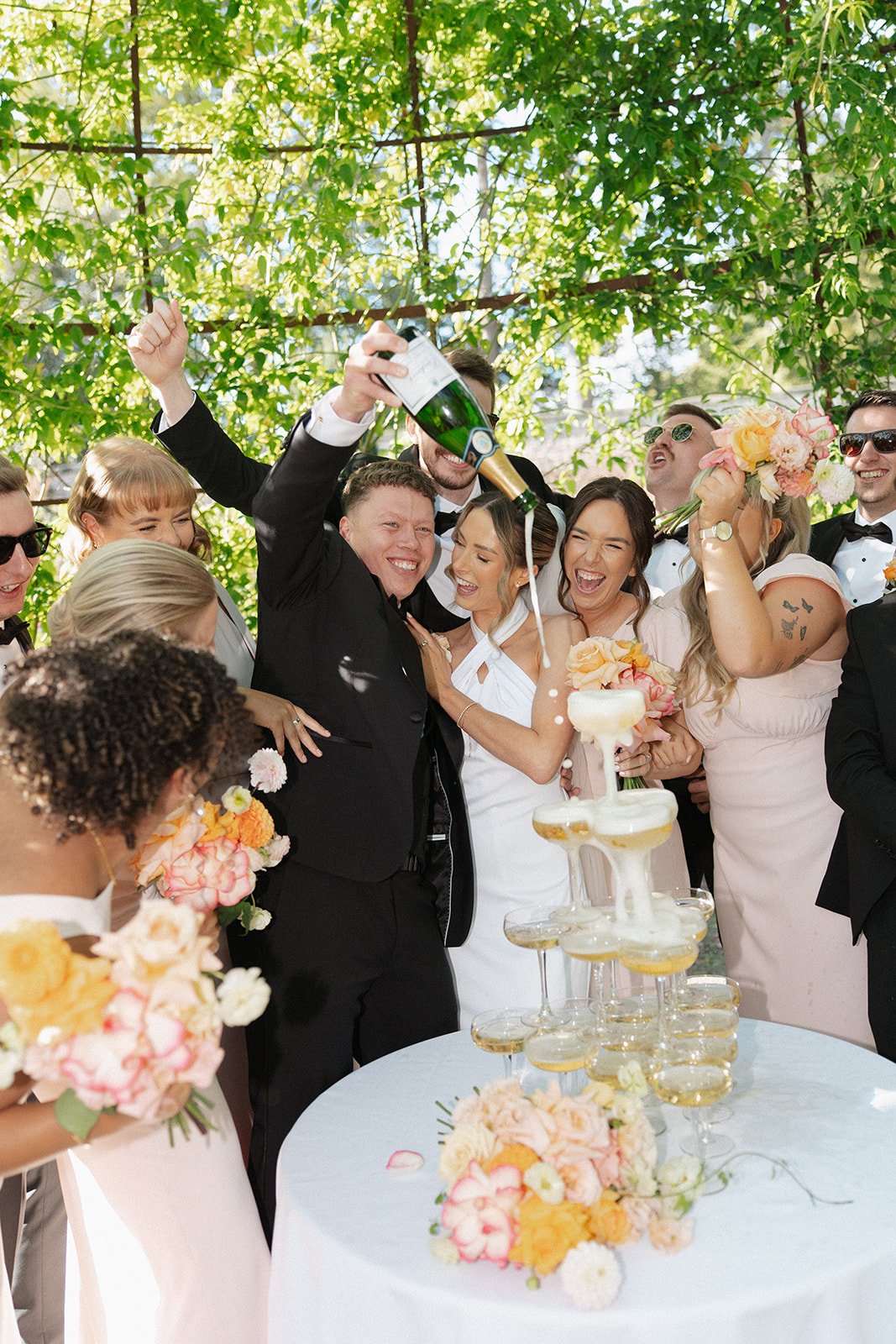 Happy wedding celebration with people pouring champagne into a tower, women holding bouquets, and smiling guests in formal attire, set outdoors with lush green trees in the background.