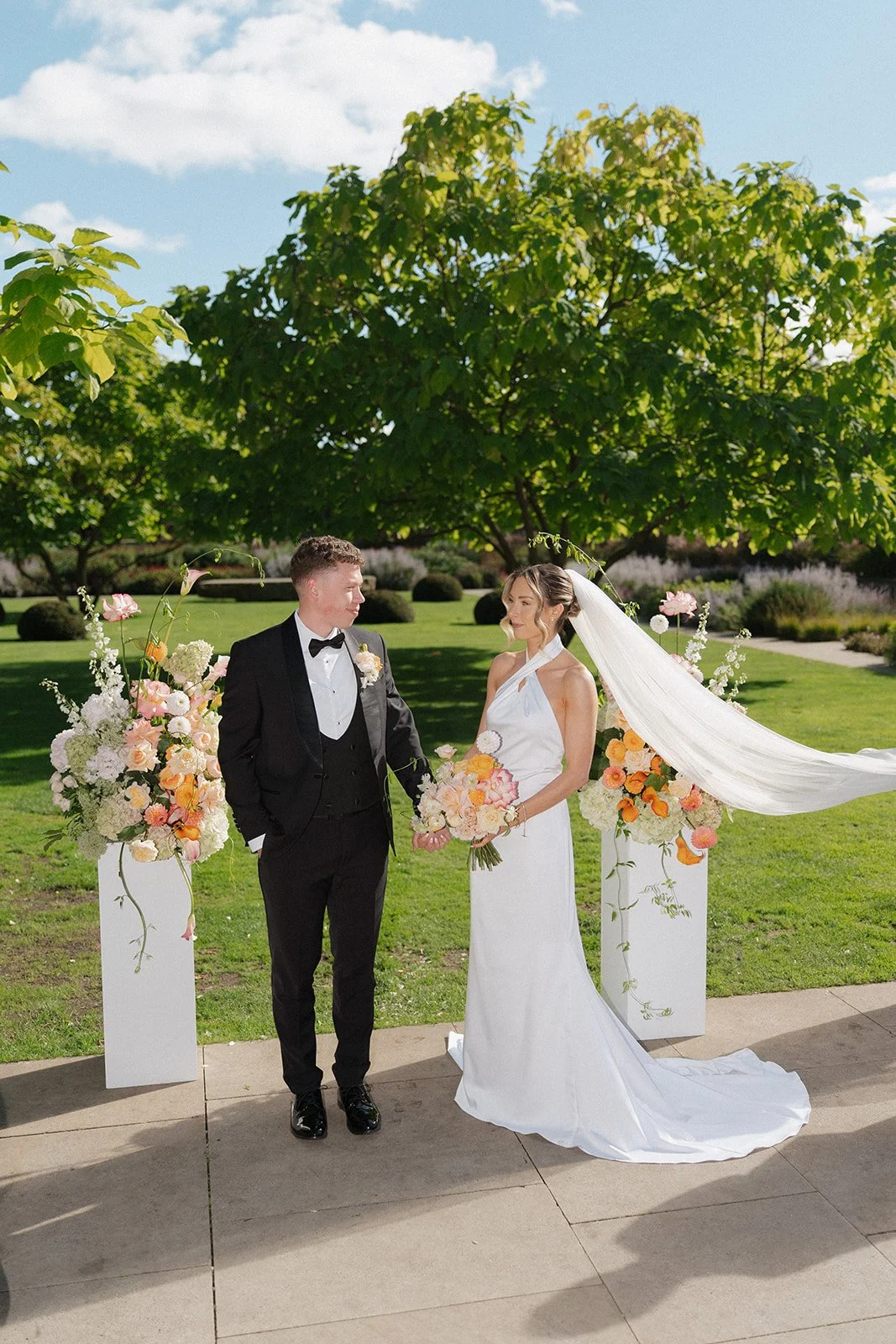 A bride and groom standing outdoors during their wedding ceremony with floral arrangements and a large tree in the background.