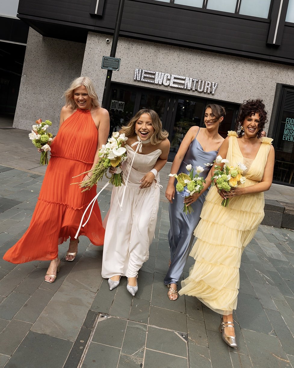 Four women dressed in colorful dresses carrying bouquets of flowers, walking cheerfully outside a modern building with a sign that reads 'New Age Century' in illuminated letters.