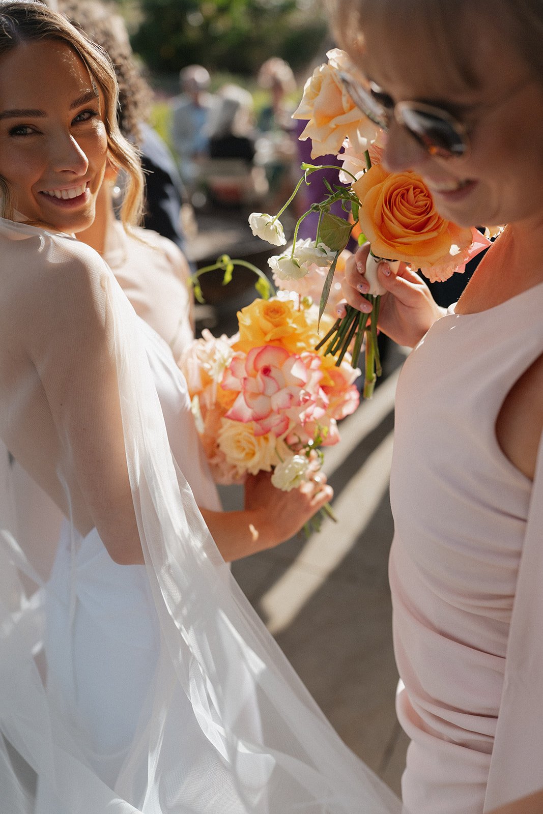 A bride smiling at another woman holding a bouquet of peach and pink roses at an outdoor wedding.