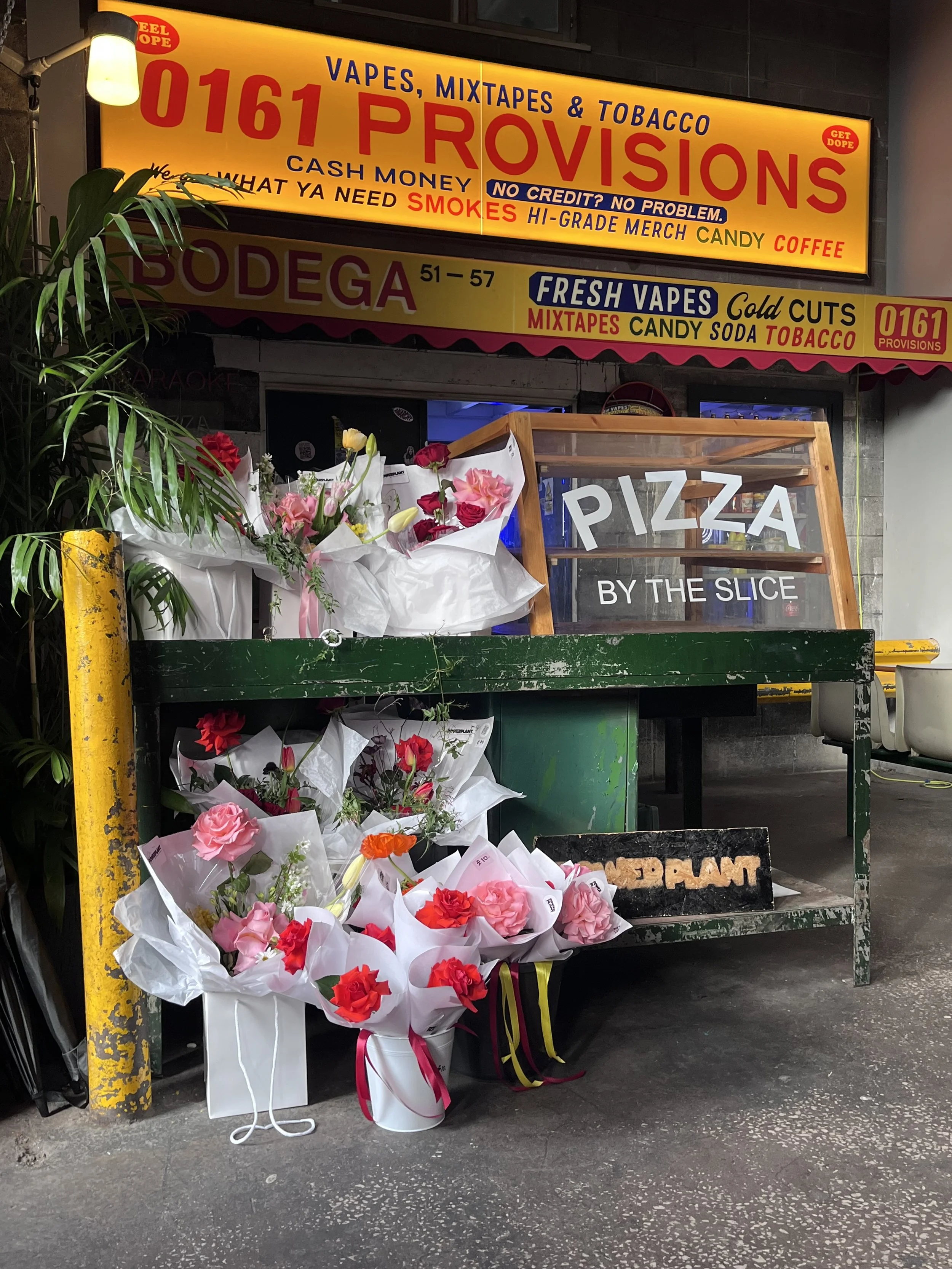 A small flower shop with bouquets of pink and red flowers on the ground and a wooden sign that says "Pizza by the Slice". Above, there are bright yellow and orange advertisements for vape and tobacco products, including the store's phone number.
