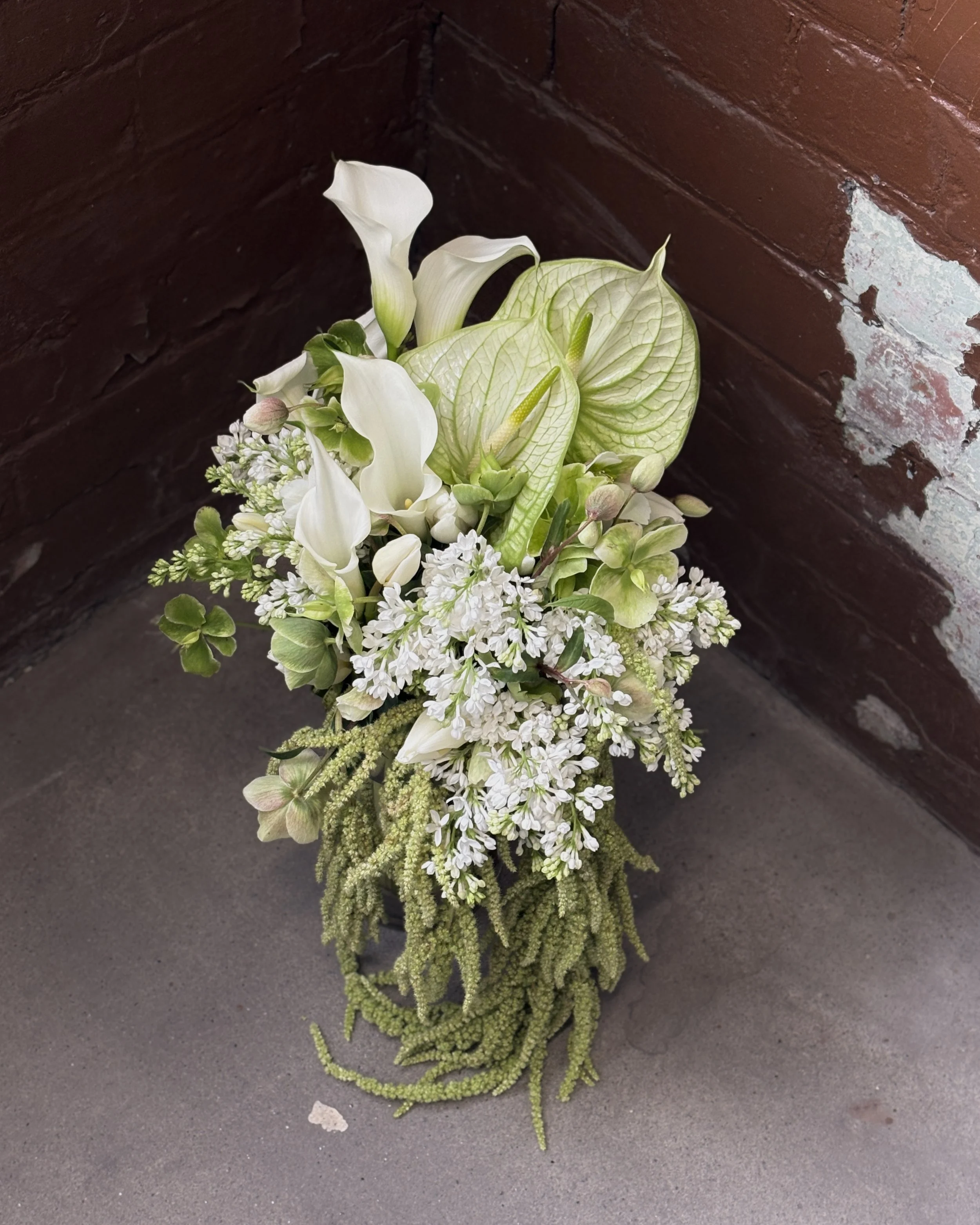 White and green flower arrangement with calla lilies, anthuriums, and other greenery, placed against a brick and concrete background.