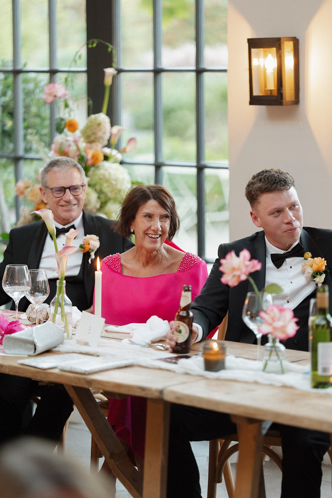People enjoying a formal celebration dinner at a decorated table with flowers, candles, and drinks in a bright room with large windows.