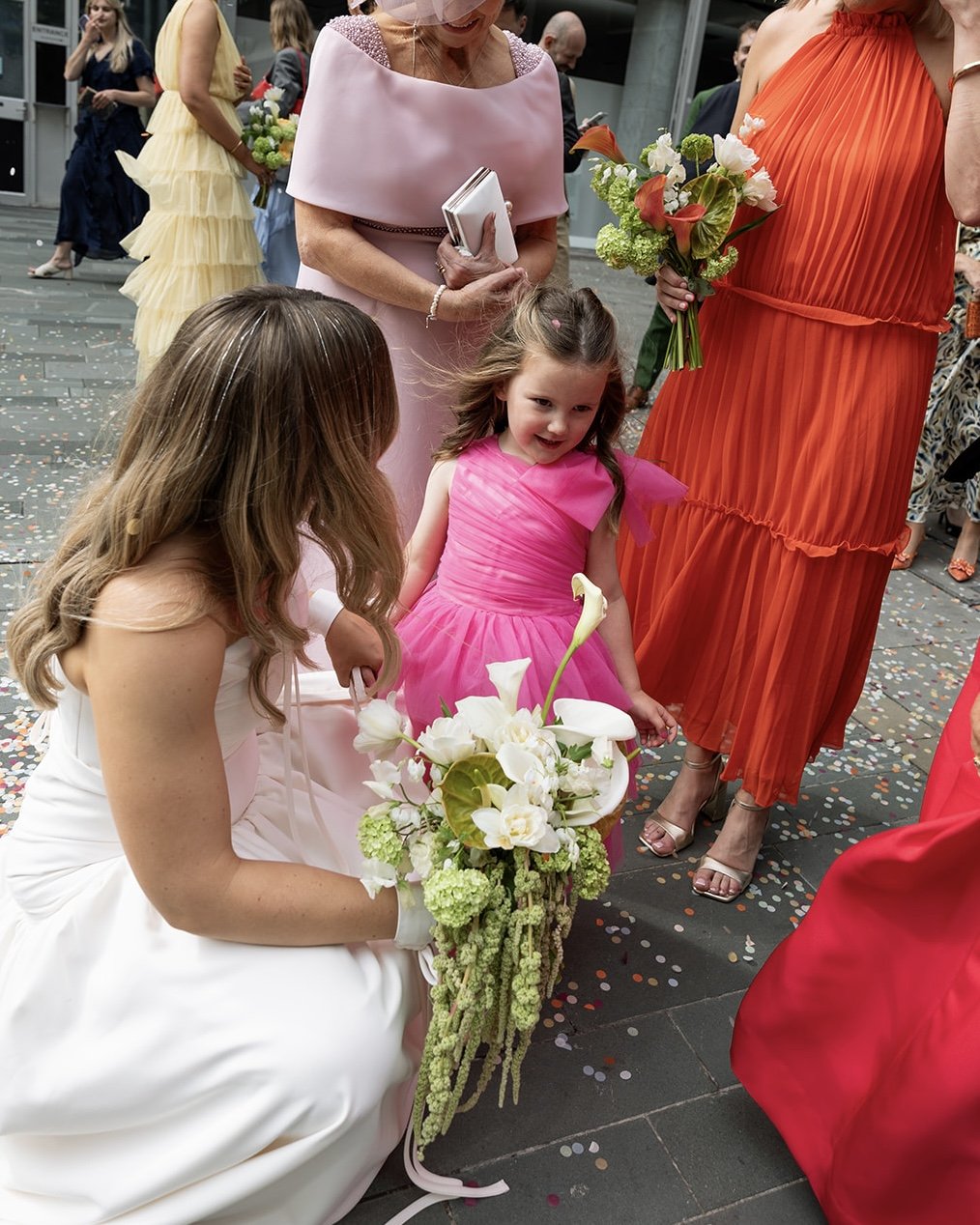 A young girl in a bright pink dress holding a bouquet of white flowers at a celebration, surrounded by women in colorful dresses and confetti on the ground.