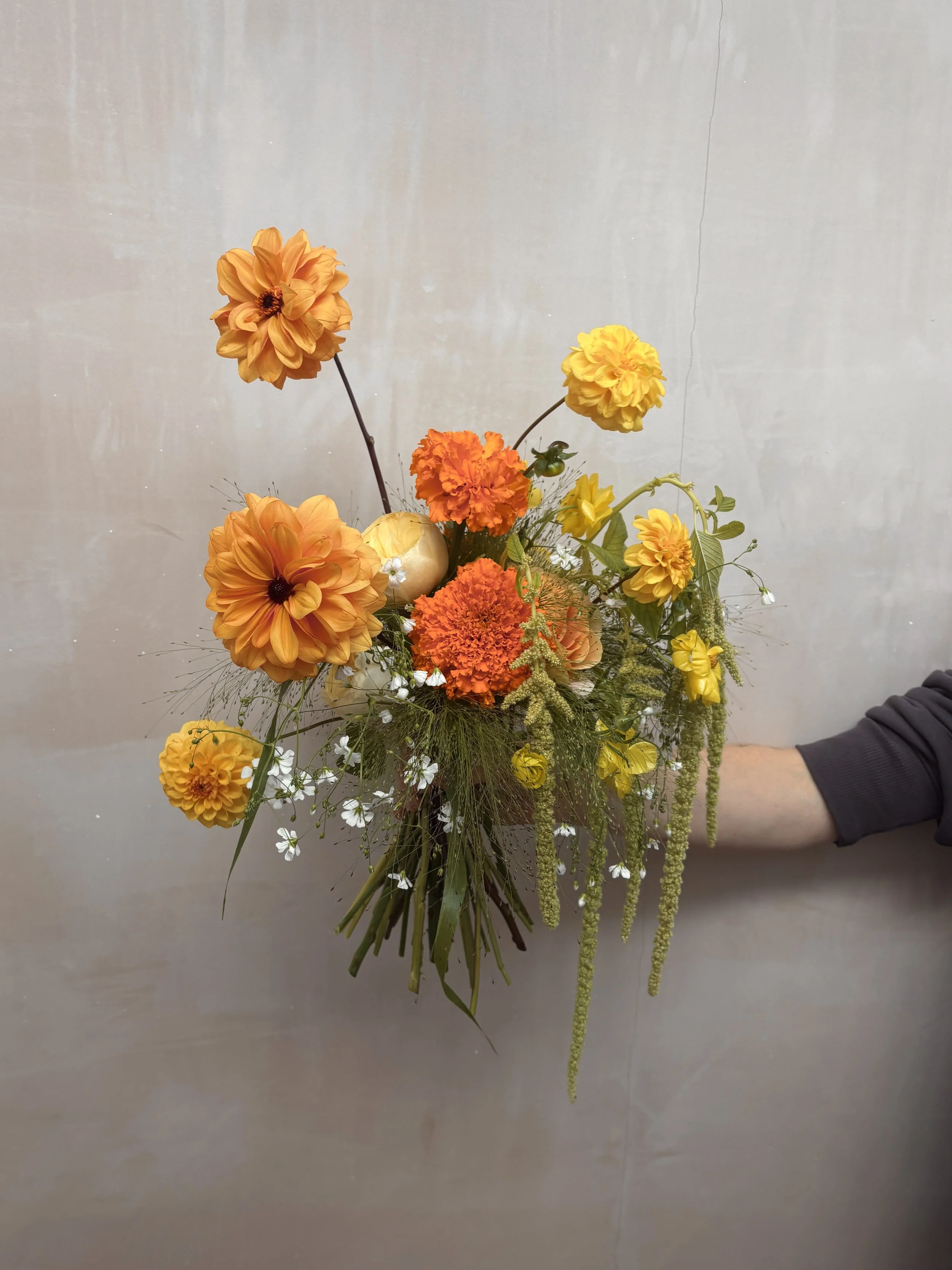 A bouquet of orange and yellow flowers with greenery, held by a person with a black long sleeve shirt against a plain neutral background.