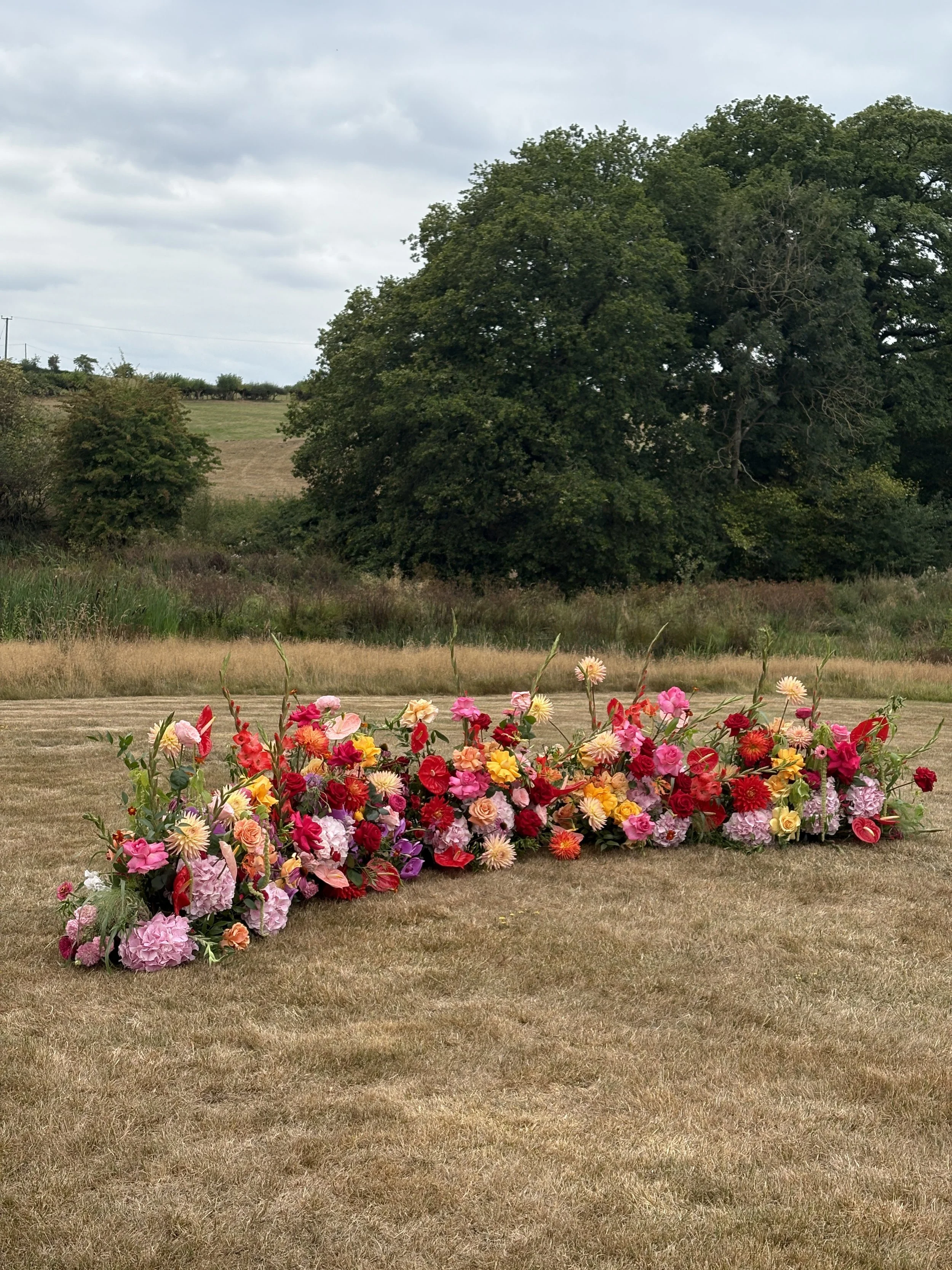 Colorful flower arrangement on a grassy field with trees and cloudy sky in the background.