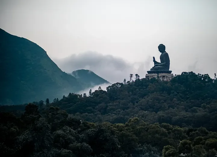 A large seated Buddha statue on a hilltop overlooking lush green trees and mountains, with overcast sky in the background.