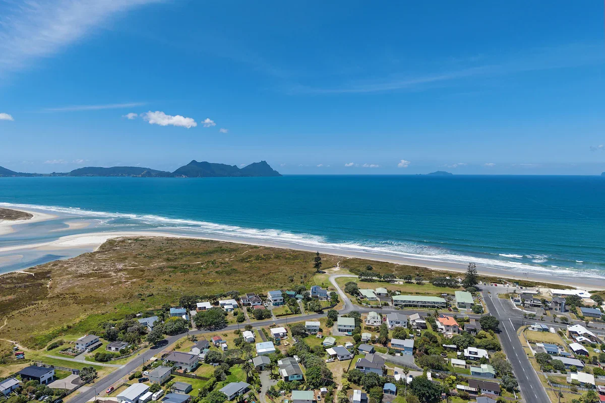 Aerial view of a coastal neighborhood with houses, roads, and green spaces next to a sandy beach and ocean, with mountains in the background and a blue sky.