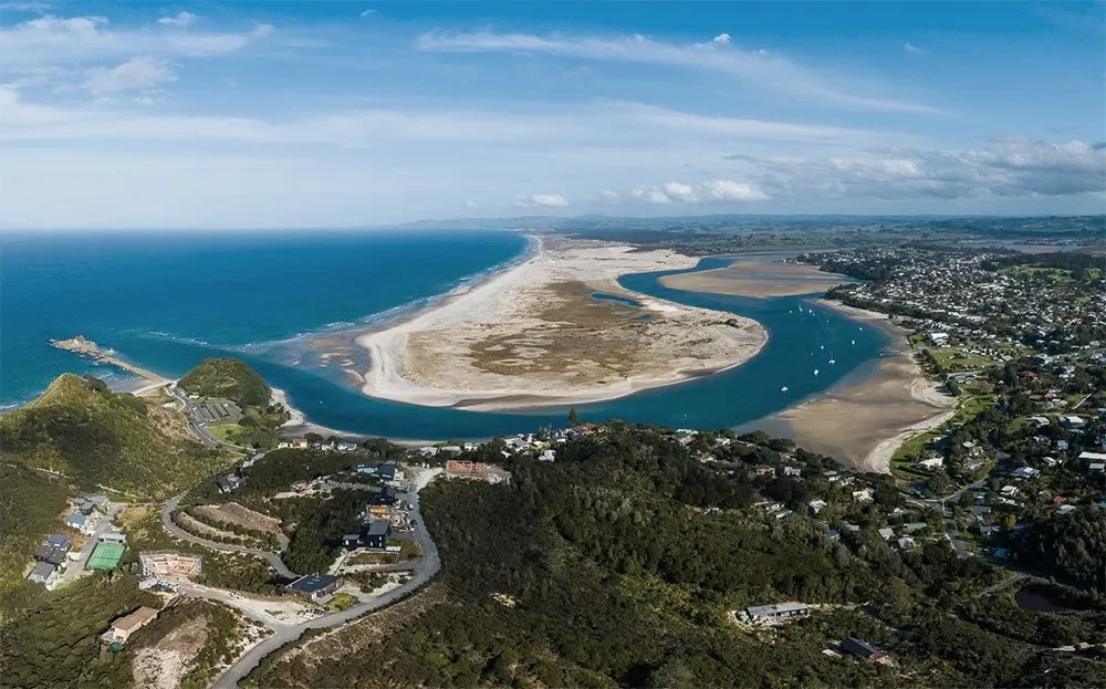 Aerial view of a coastal town with a winding river separating land areas, houses and green hills, boats on the water, sandy beaches, and the ocean in the distance under a partly cloudy sky.