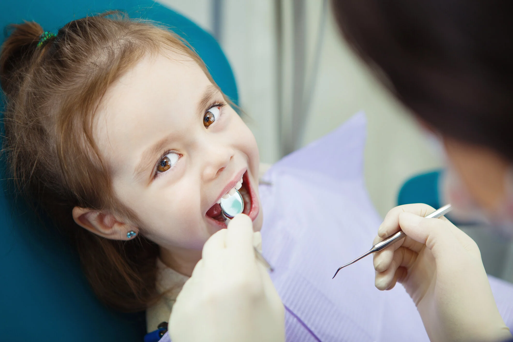 Young girl with brown hair and earrings smiling at the dentist while getting her teeth examined with dental tools.