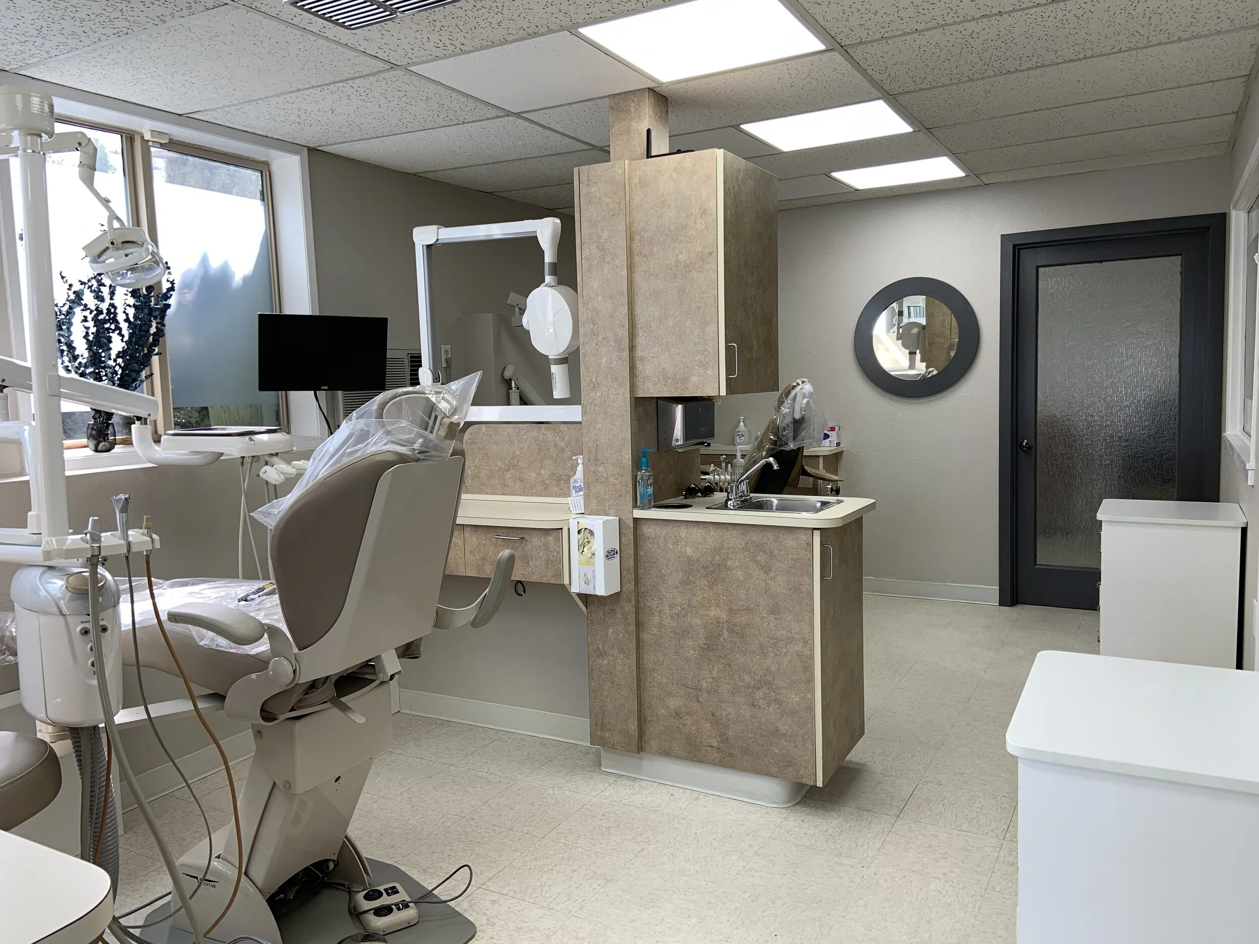 A clean, empty dental examination room with dental chair, equipment, and a small sink area.
