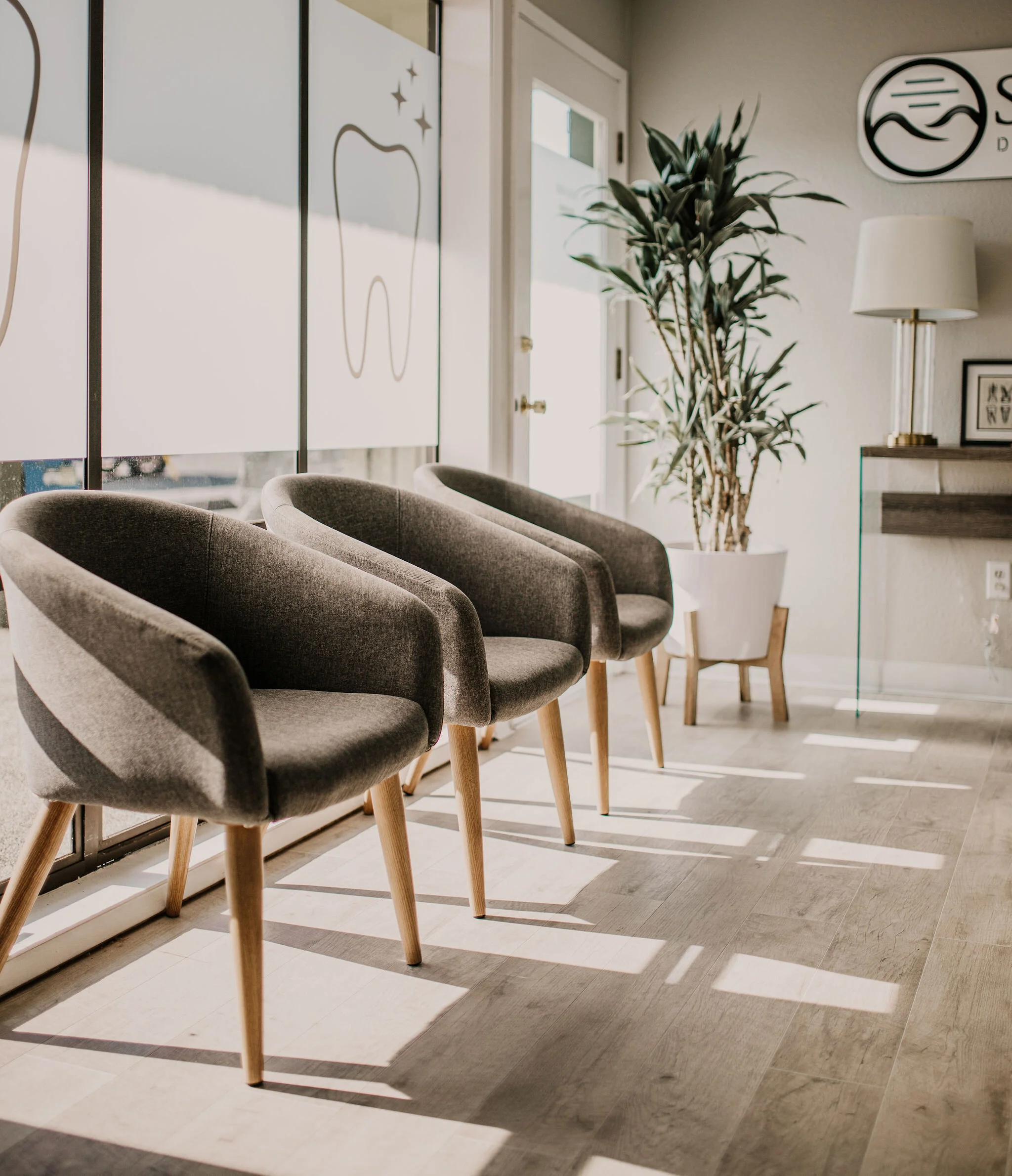 Interior of a waiting room with four gray upholstered chairs with wooden legs, a potted plant, a white lamp, and framed artwork on the wall, illuminated by natural light.