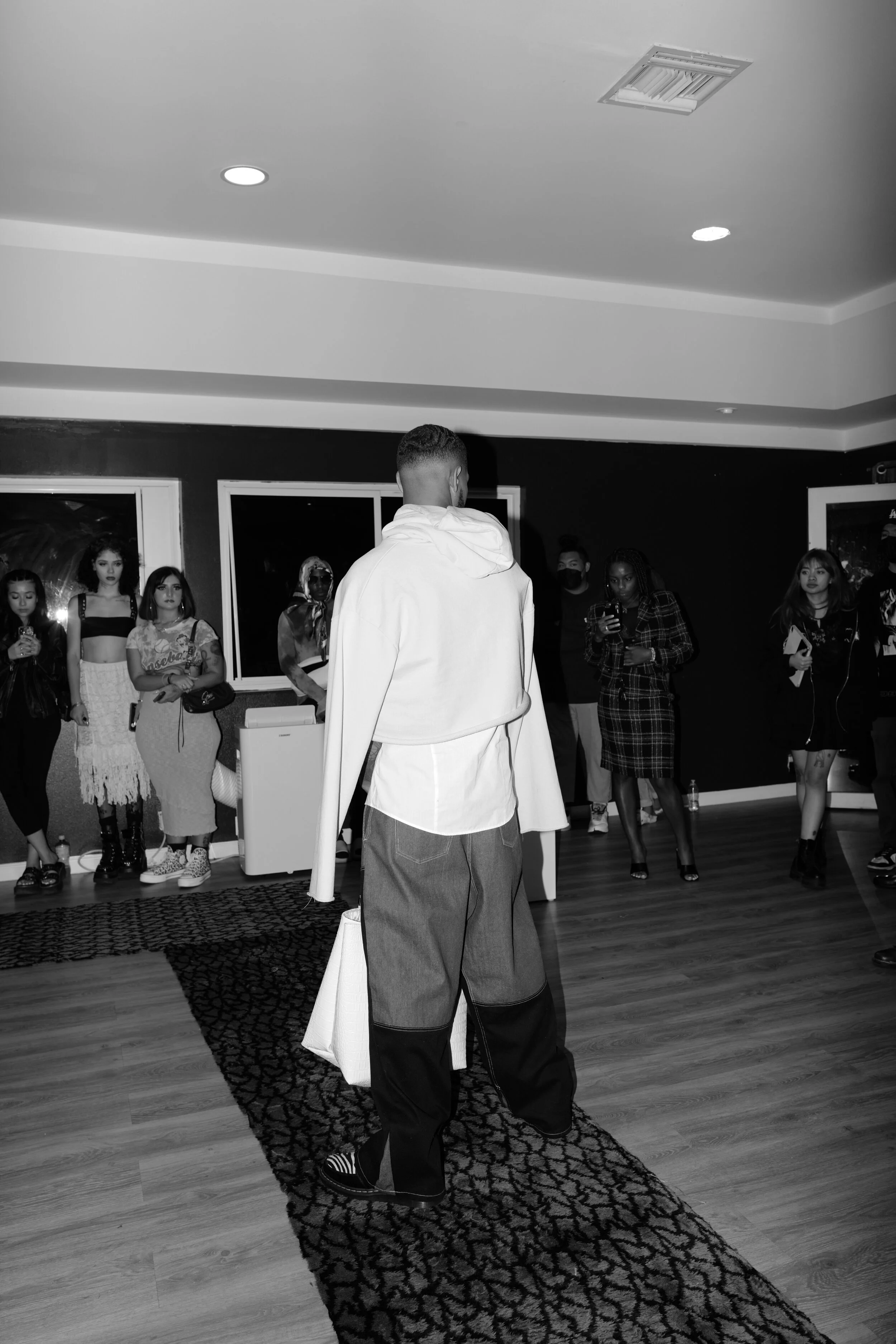 A group of young adults gathered in a room with dark walls and large windows, listening to a man with short hair and wearing a white hoodie and baggy pants, who stands with his back to the camera. The group includes women with varied hairstyles and outfits, some holding drinks or phones, and appears to be engaging in a social event.