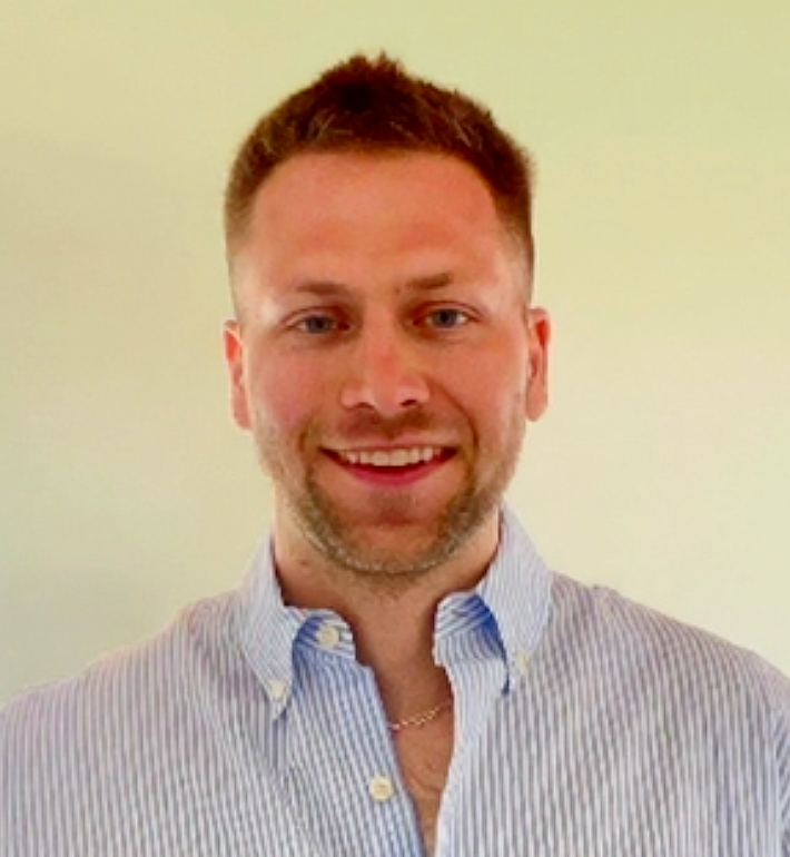 A smiling man with short brown hair, a light beard, wearing a blue striped button-up shirt and a necklace, standing in front of a plain light-colored wall.