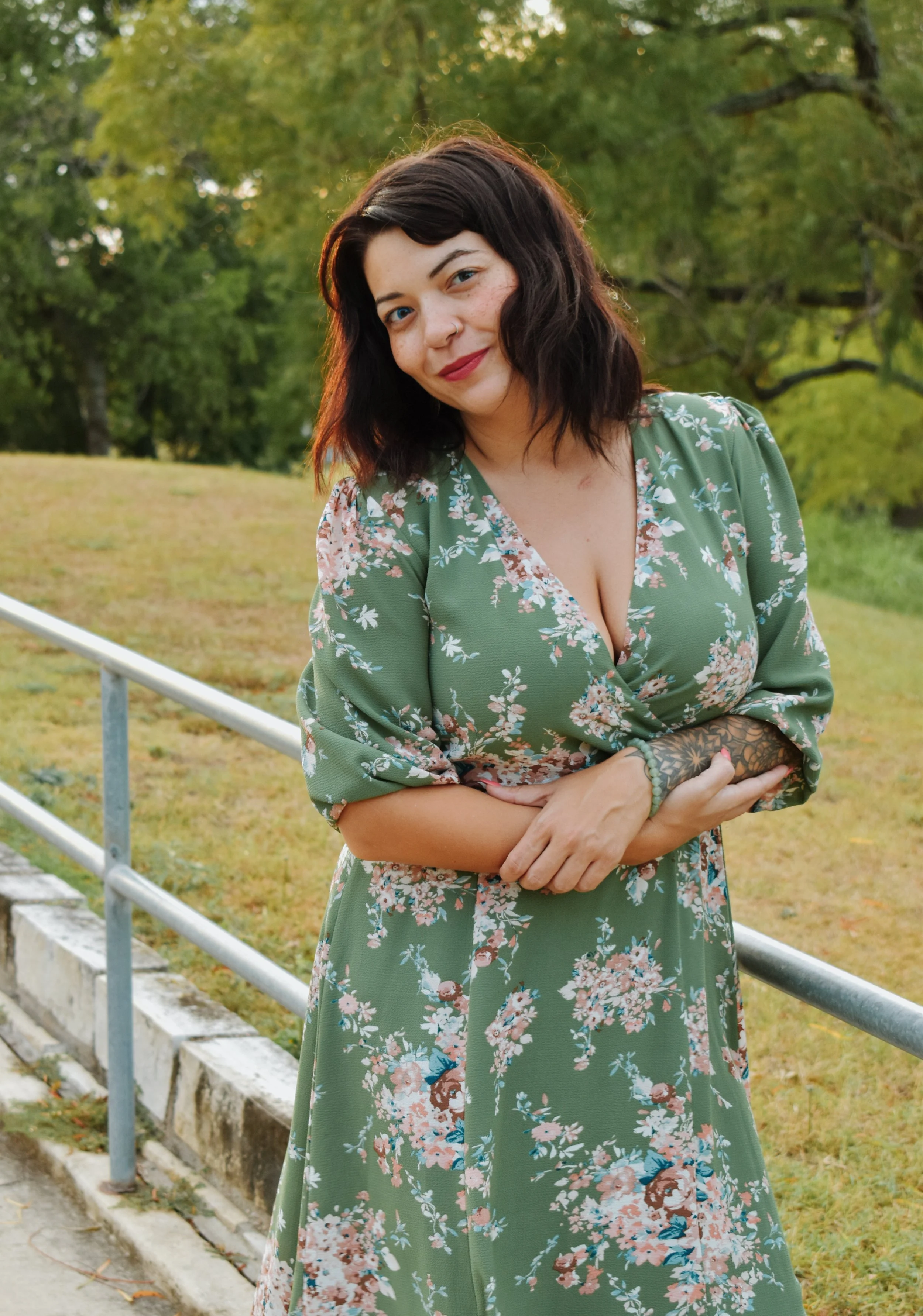 A woman with dark hair and fair skin standing outdoors near a park railing, wearing a green floral dress and smiling.