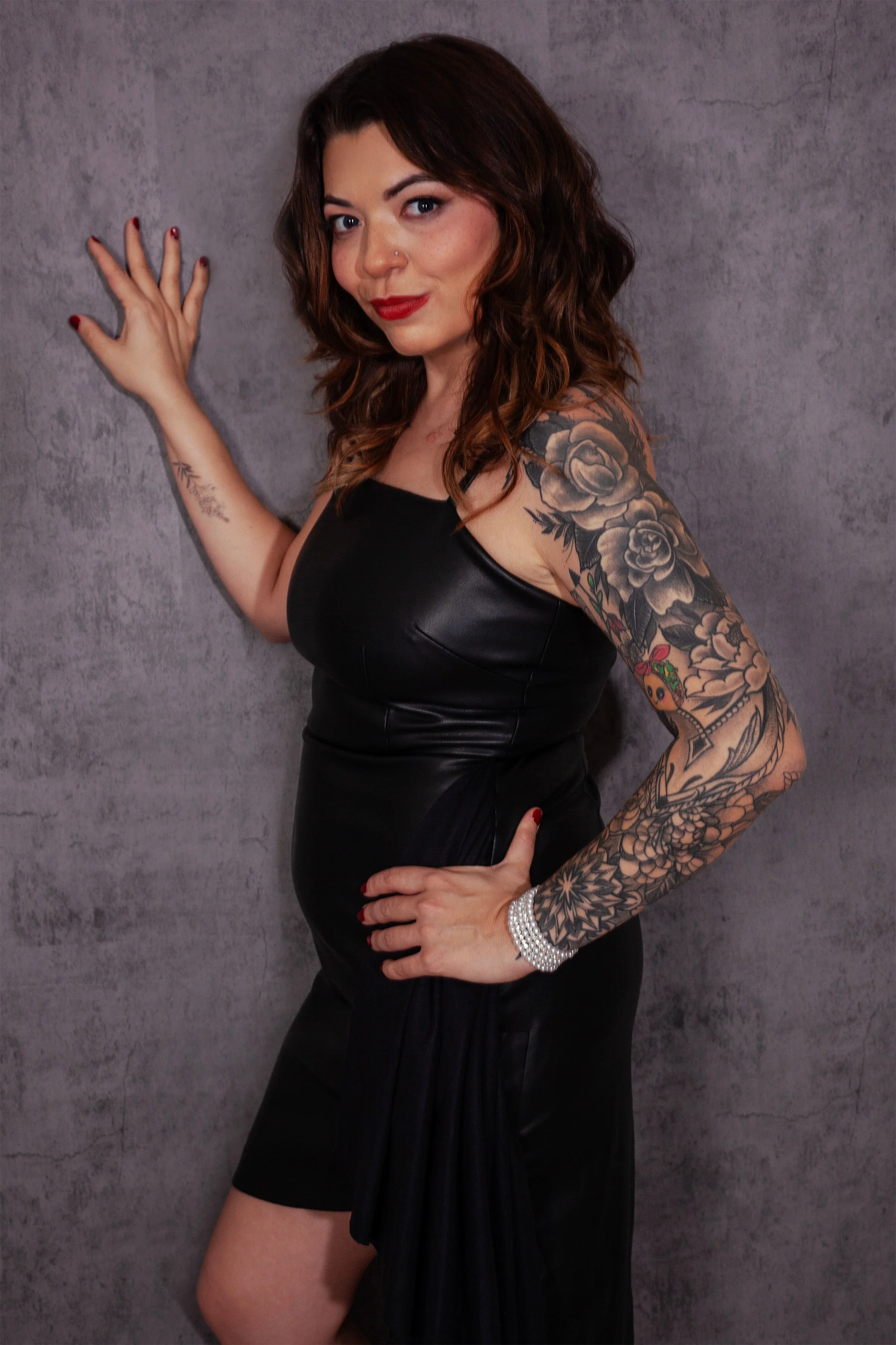 A woman with dark brown hair, red lipstick, and tattoos on her left arm, wearing a black dress, standing against a gray textured wall, posing confidently with her left hand on her hip and her right hand touching the wall.