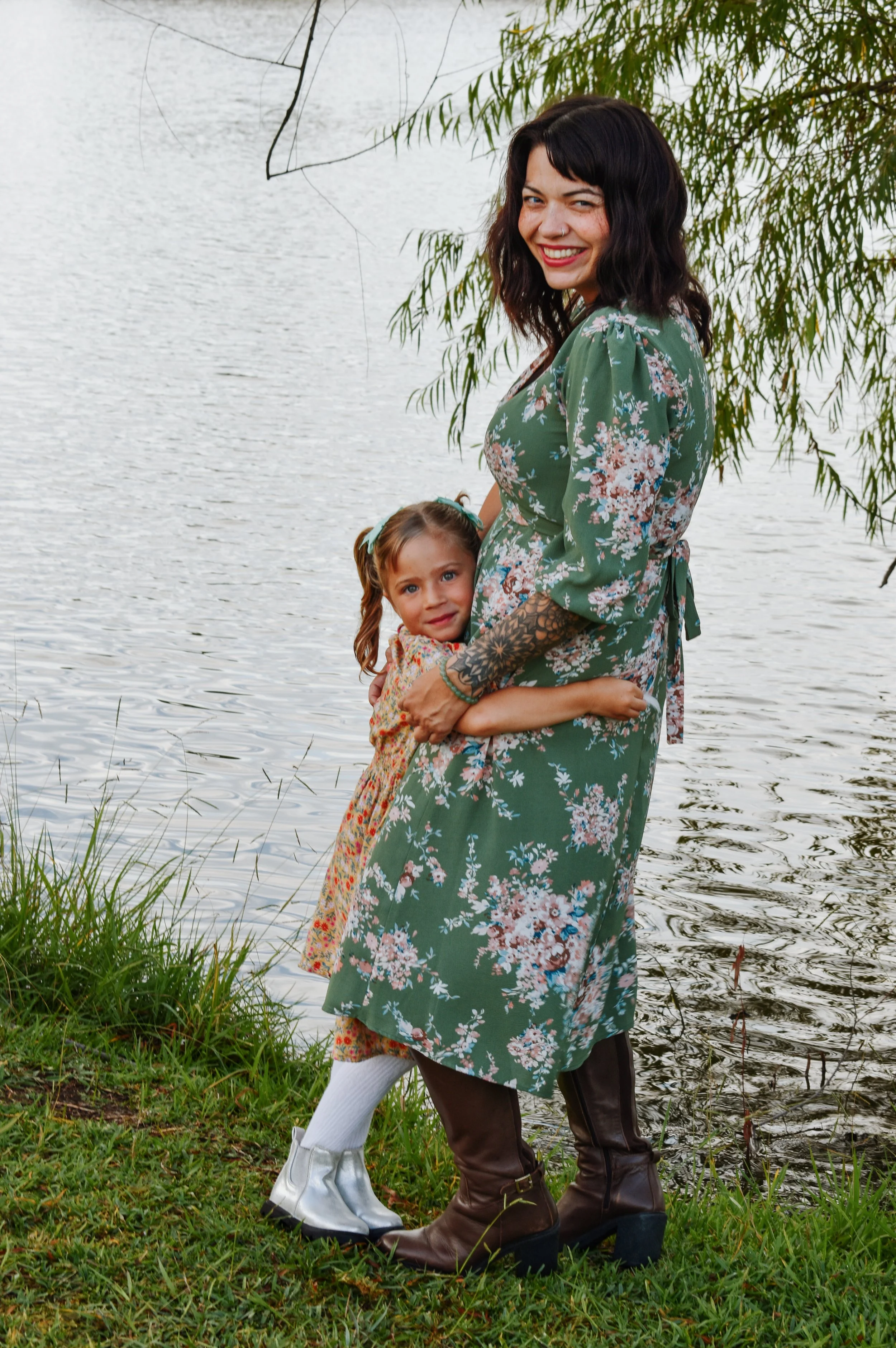 A woman and a young girl hugging near a body of water with trees in the background. The woman has dark, wavy hair and tattoos on her arm, wearing a green floral dress and brown boots. The girl has light brown hair, wearing a patterned dress and silve