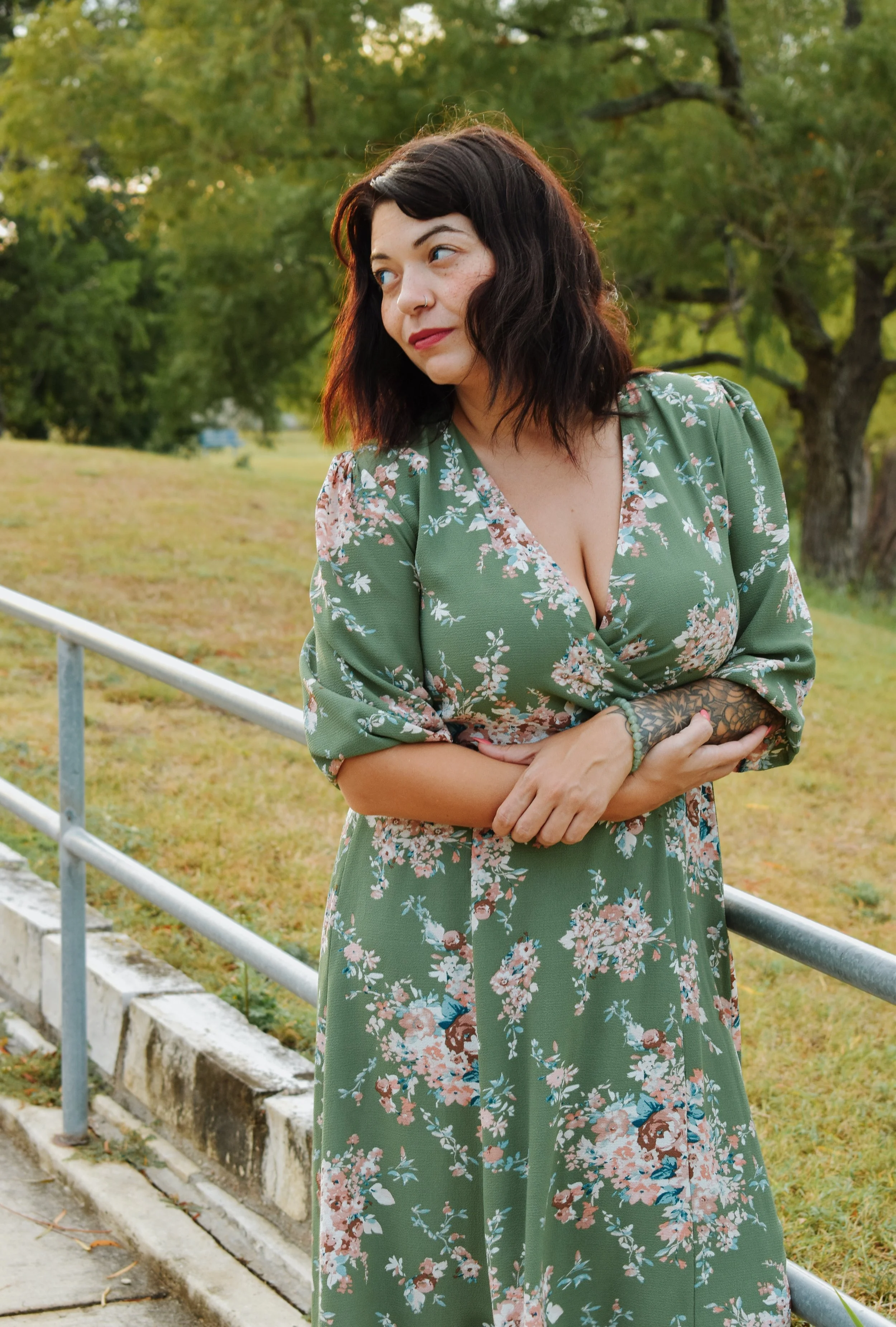 A woman with dark hair and tattoos on her arms, wearing a green floral dress, standing outdoors near a metal railing, with trees and grass in the background.