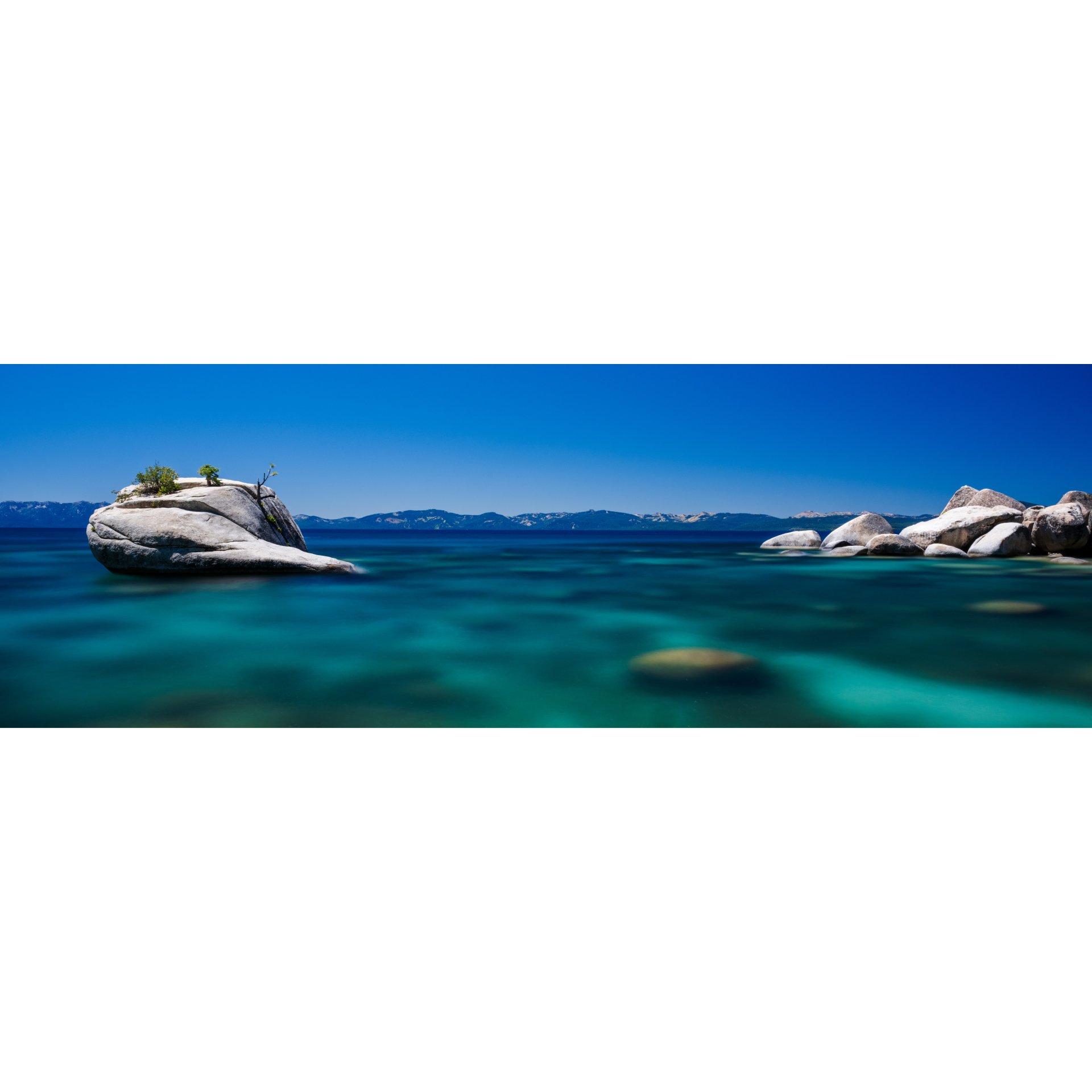Large rock in clear blue water with distant mountains.