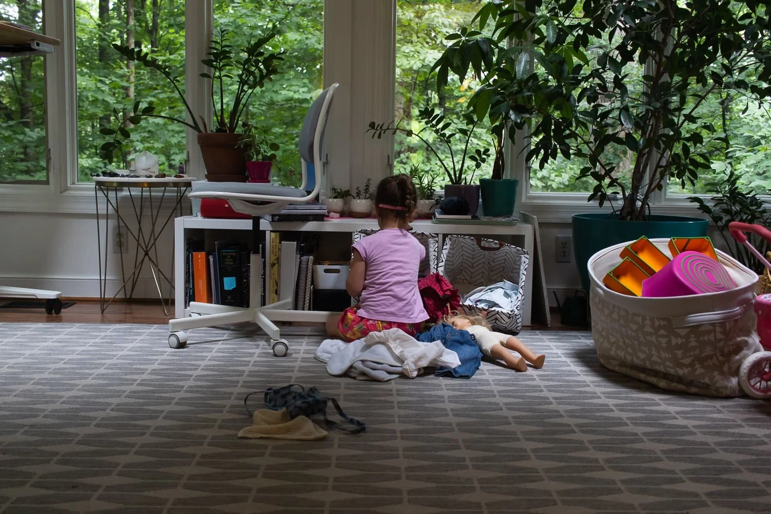 A young girl sitting on the floor in a room filled with indoor plants, surrounded by various household items and toys, with large windows showing a lush green outdoor scene.