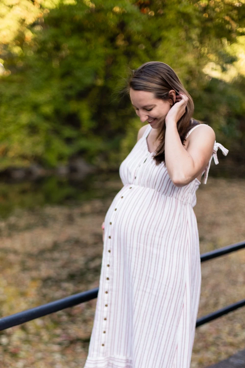A pregnant woman smiling while looking down, outdoors with blurred green trees in the background, wearing a sleeveless, striped dress.