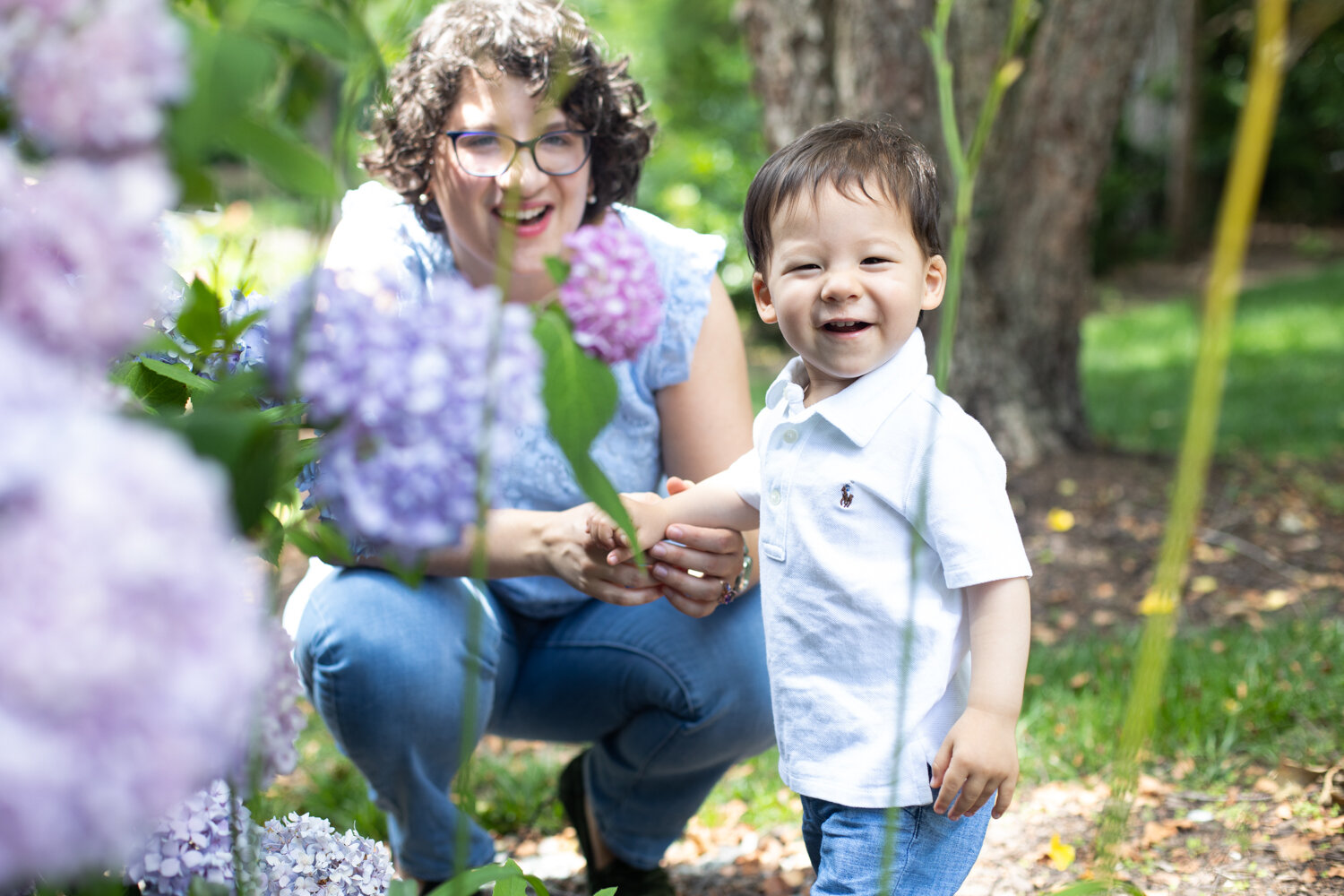 Little boy squinty smile with mom at purple hydrangea bush in backyard