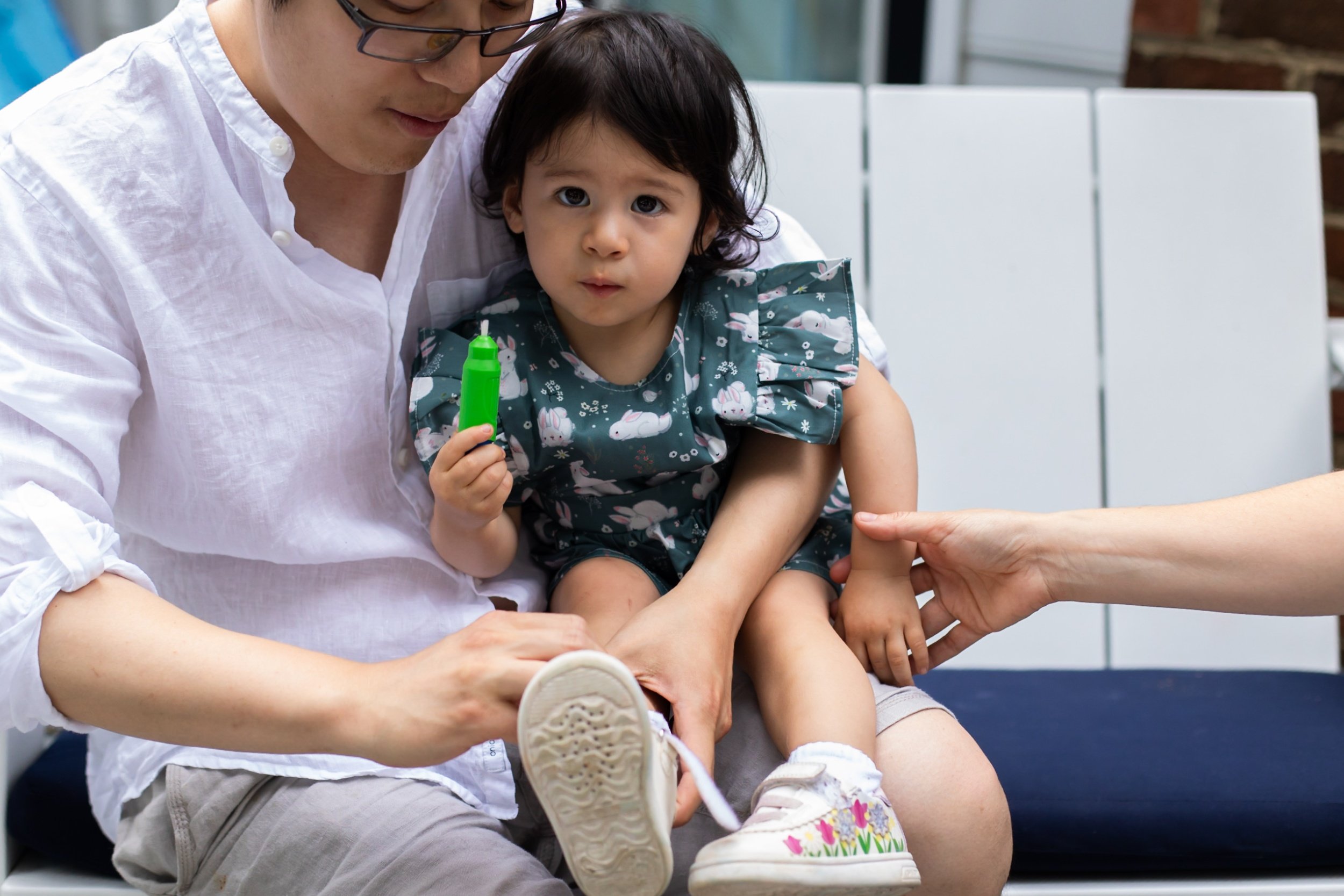 A little girl holds a paint brush and stares at the camera in her dad's lap while he puts on her shoes. Mom's hand is lightly holding her other arm.