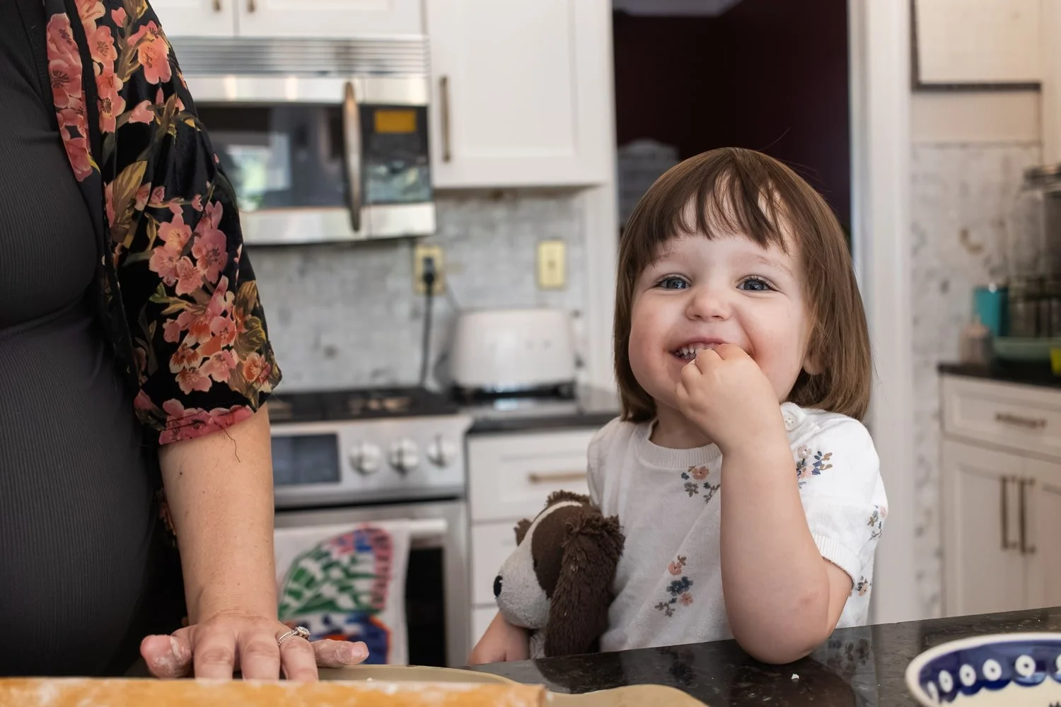 A young girl with brown hair and a floral shirt licks her finger while standing at a kitchen counter. Her pregnant mother, partially visible, is rolling out pizza dough on a wooden board.