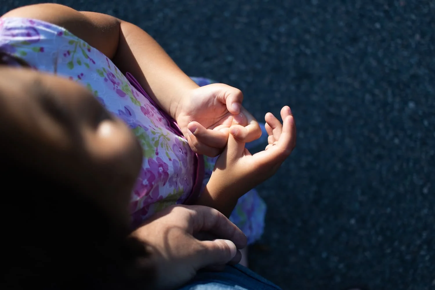 A child's hands resting on an adult's hands, with the child's fingers intertwined and the adult's hand gently holding the child's wrist, on a dark outdoor surface.