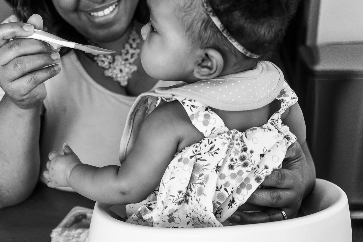 A mom smiles while feeding her baby daughter apple sauce and supporting her daughter's back as she sits on the table.