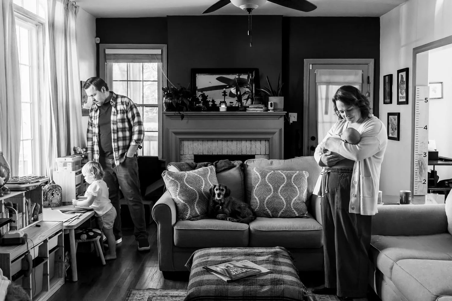 Black and white image of a family of four in their living room. Their dog is the only one looking at the camera, as she sits on the couch. Mom holds her newborn daughter and smiles down at her while Dad and toddler daughter play near the window.