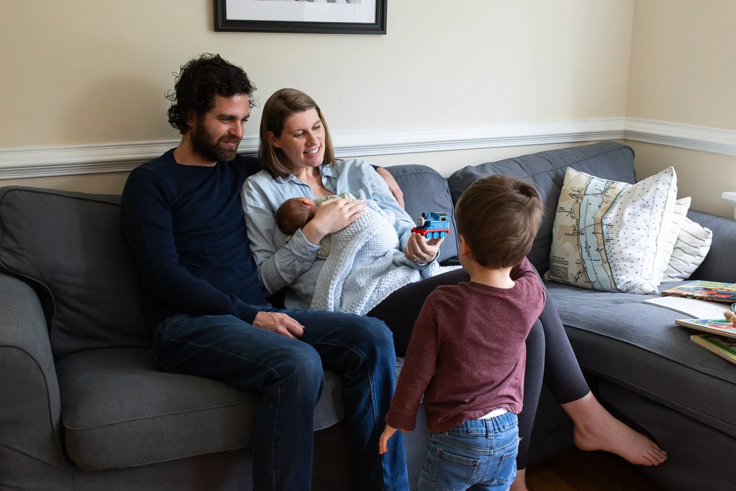 Family of 4 in their living room. Mom is holding baby and showing a toy to her toddler while Dad sits close and smiles at them.