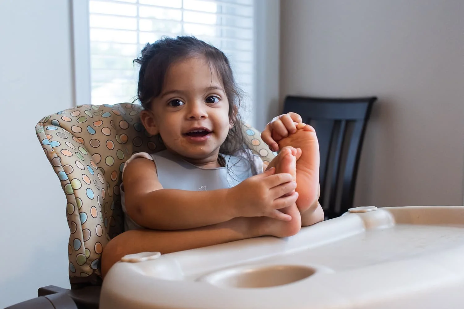 A toddler sitting in her high chair, her face covered in food. She has pulled her feet up above the tray and is playing with her toes while she smiles broadly at the camera.