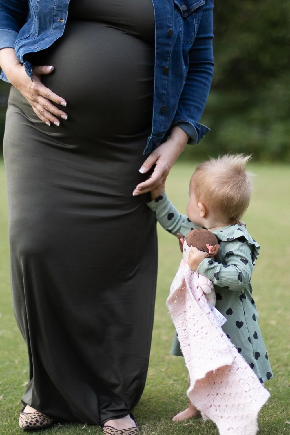A pregnant woman standing outdoors with her hand on her belly, wearing a long black dress and a denim jacket. A small child standing beside her, holding a plush toy and touching her belly, dressed in a green outfit with black heart patterns. The back