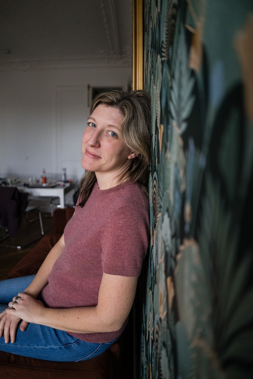 A woman with light brown hair and fair skin, dressed in a pink t-shirt and blue jeans, sitting on a chair against a colorful leafy-patterned wall inside a room. She looks relaxed and is smiling softly at the camera.