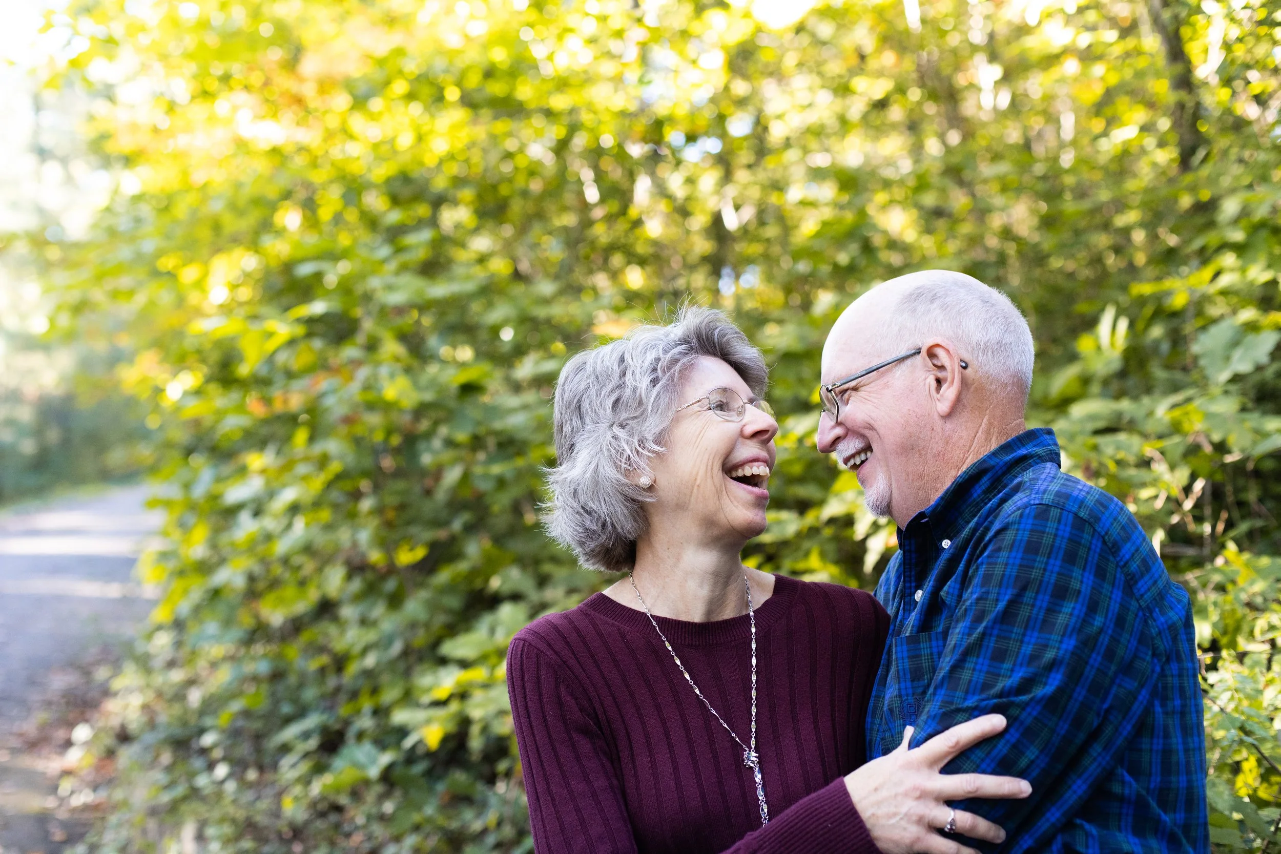 An older couple look at each other and laugh, standing in front of green foliage in a park in Richmond, VA.