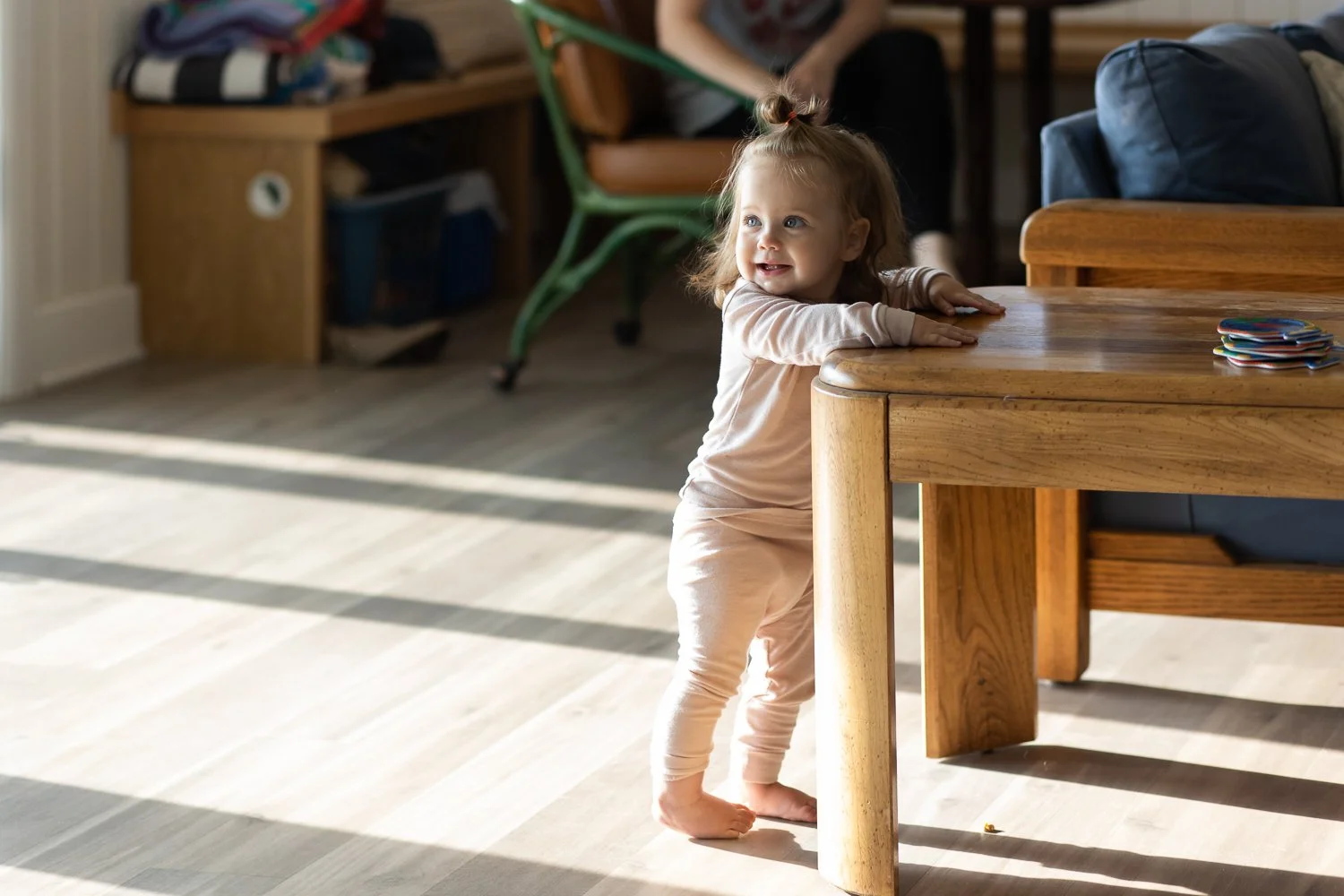 A young toddler stands beside an end table in the sunlight at home. She is smiling off camera in her pajamas.