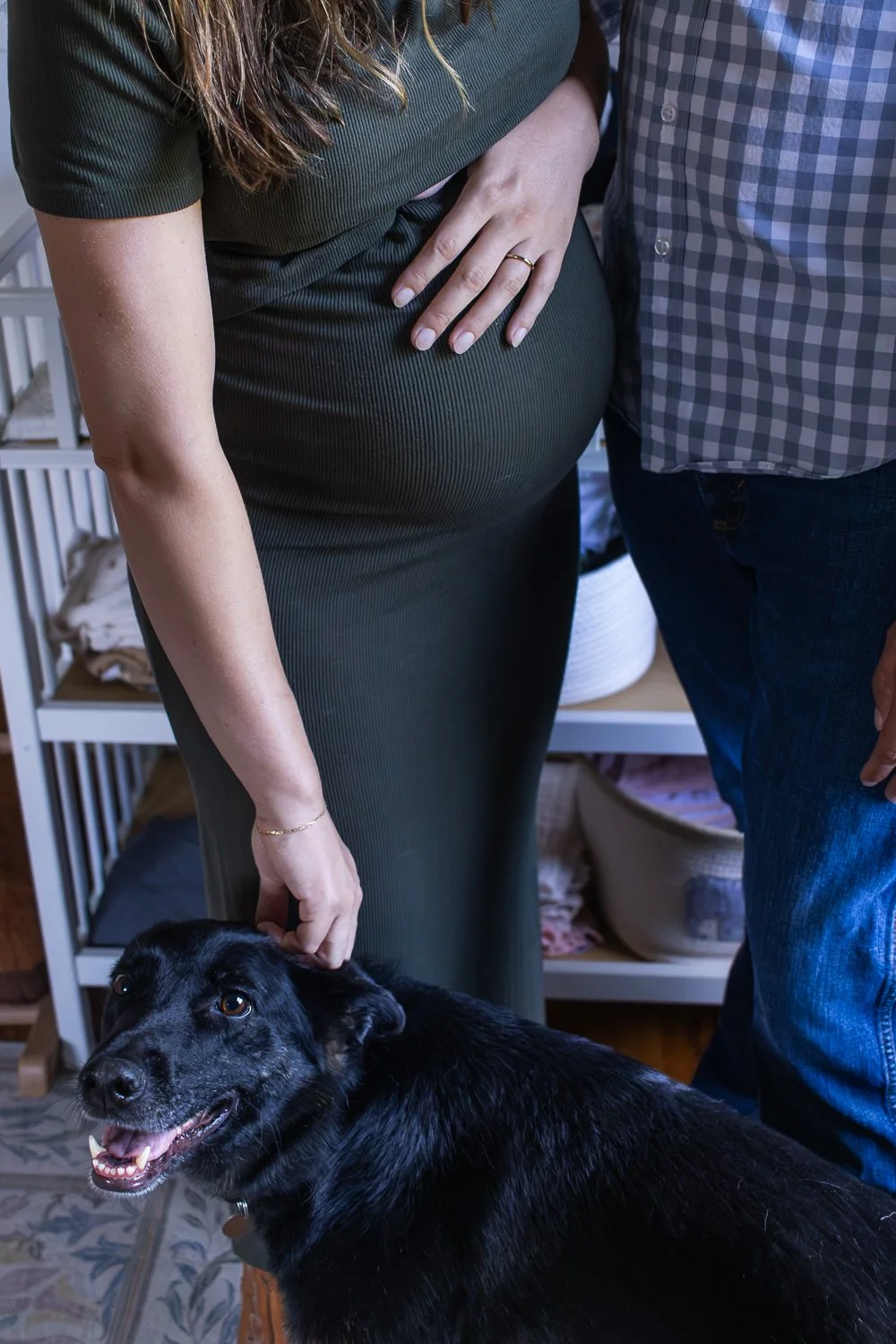 A pregnant woman standing next to a man, with her hand resting on her belly, while a black dog looks up at the camera and her other hand gently touches the dog's head.